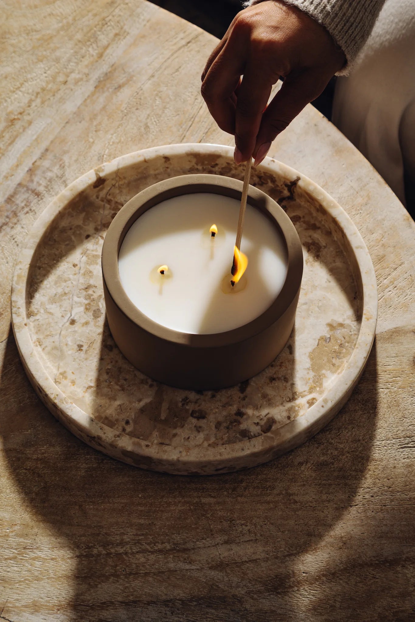 Hand lighting Rituale three-wick concrete candle with a match on marble tray