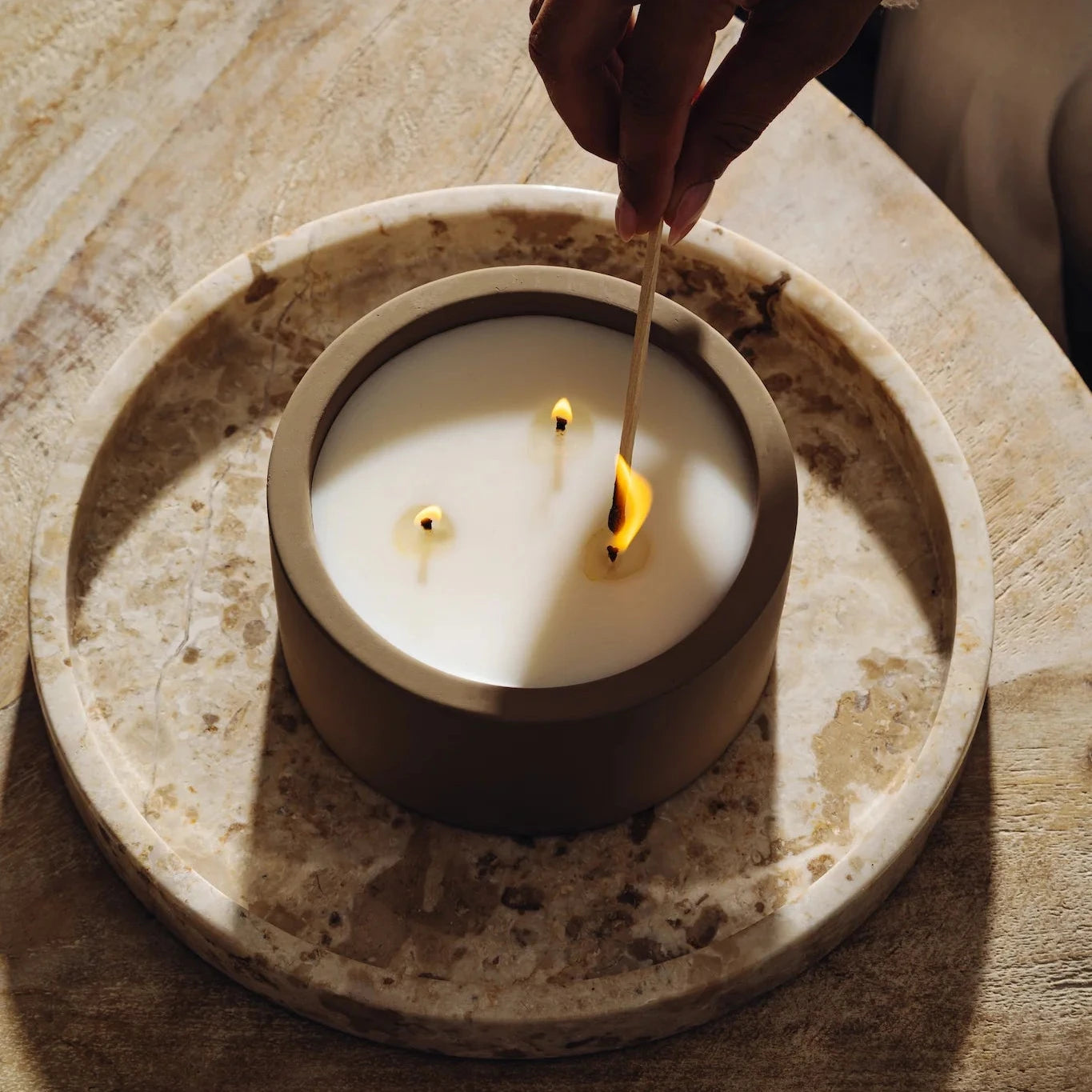 Hand lighting Rituale three-wick concrete candle with a match on marble tray
