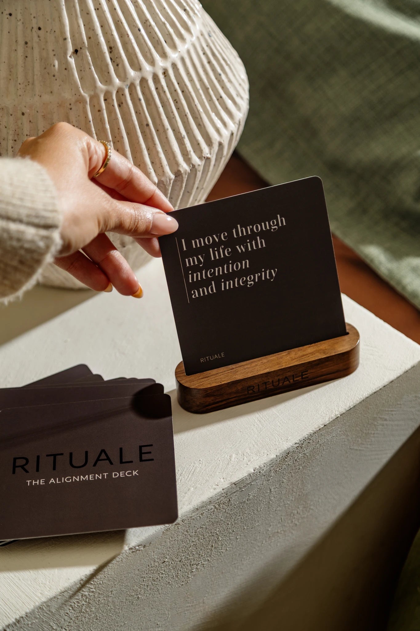 Hand holding affirmation card from Rituale Alignment Deck displayed in wooden stand on bedside table