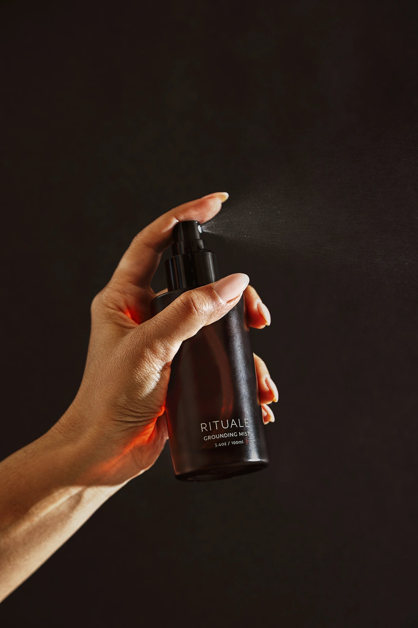 Hand holding and spraying Rituale Grounding Mist bottle against dark background