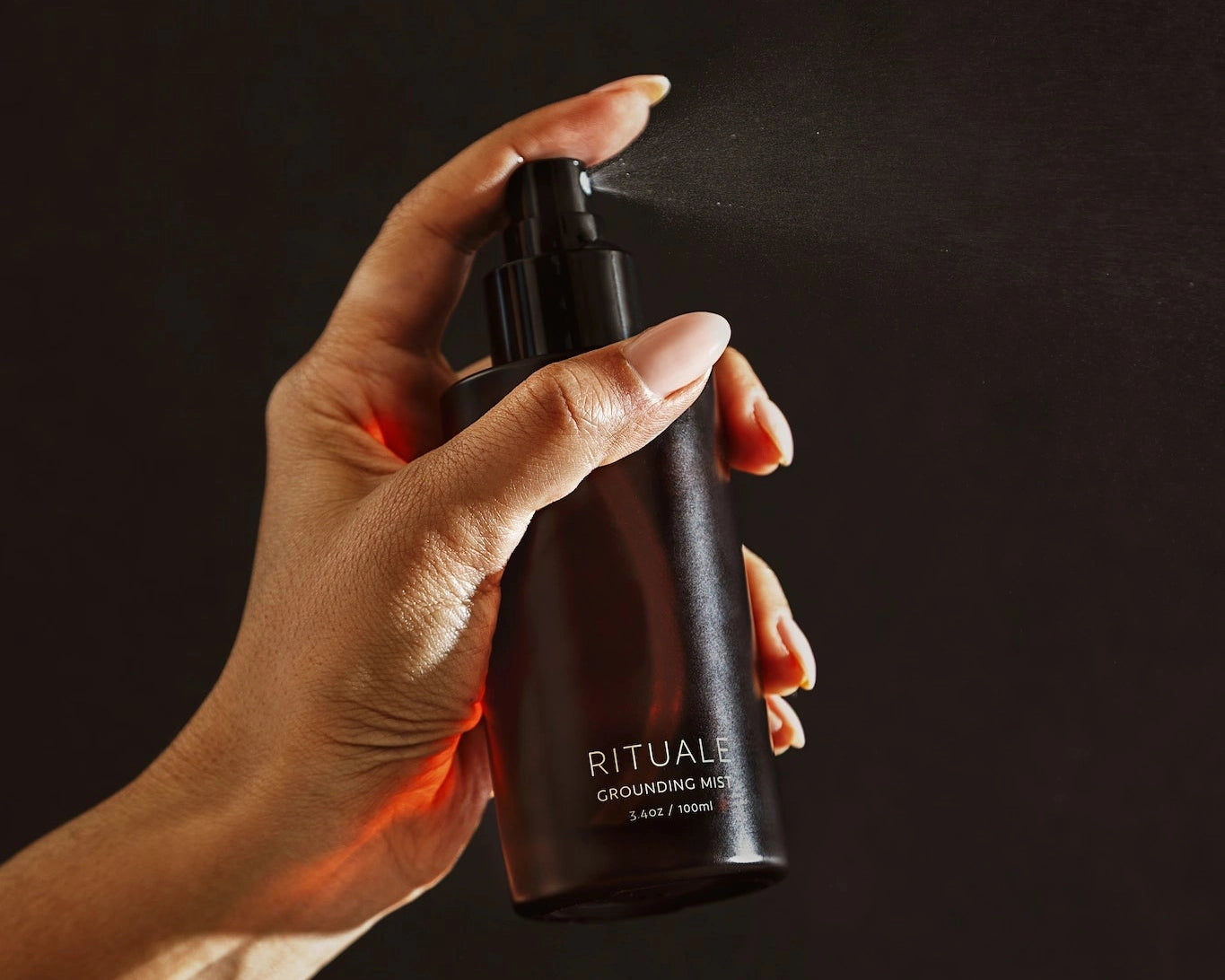 Hand holding and spraying Rituale Grounding Mist bottle against dark background