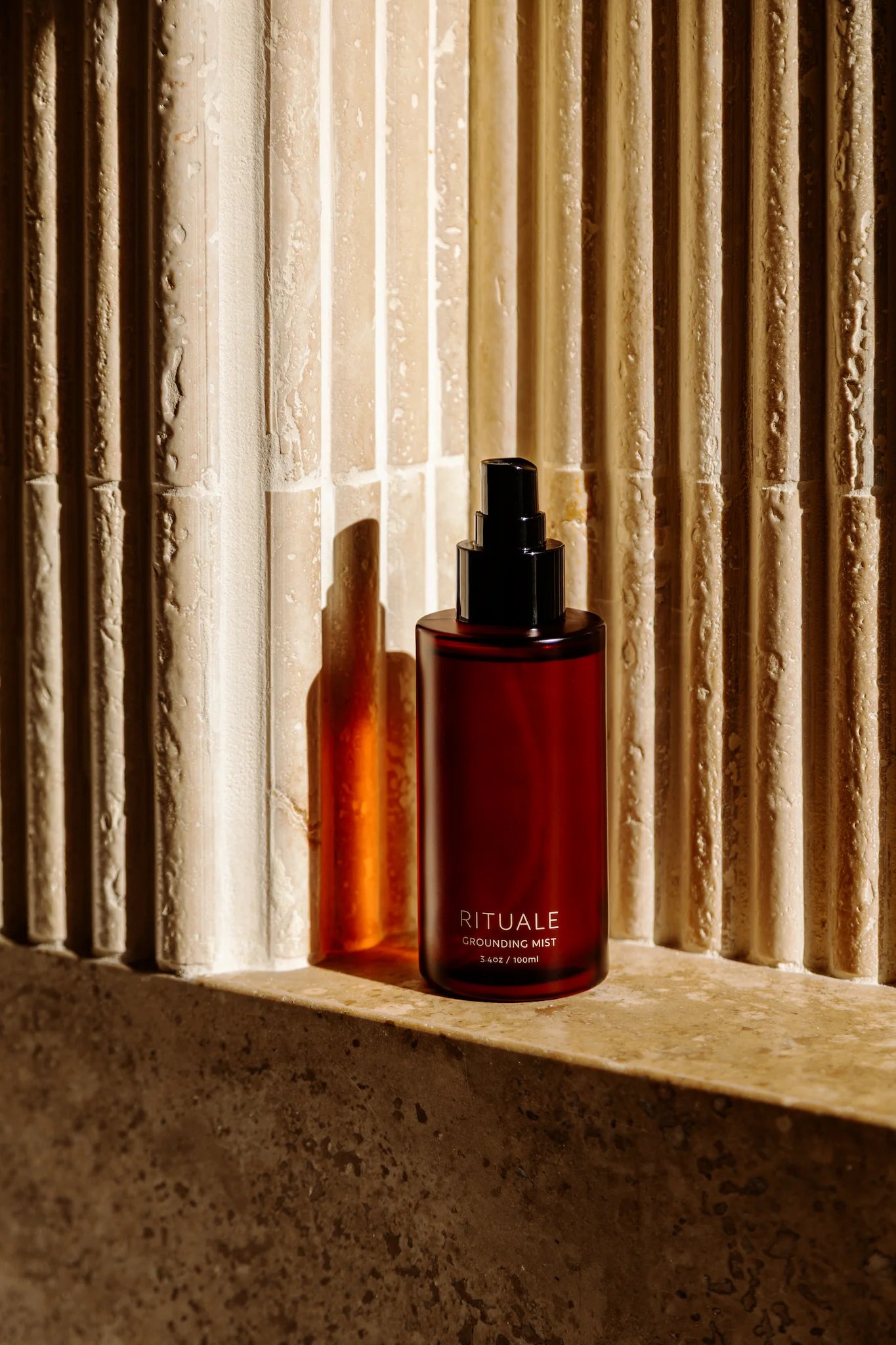 Rituale Grounding Mist amber bottle on stone ledge with warm light and fluted travertine wall background