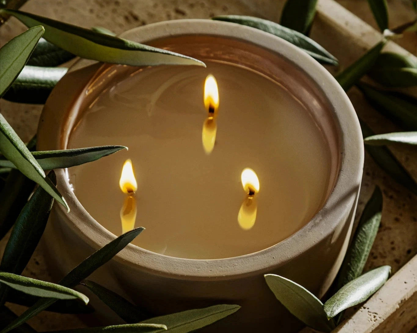 Rituale three-wick concrete candle burning surrounded by olive branches on travertine tray