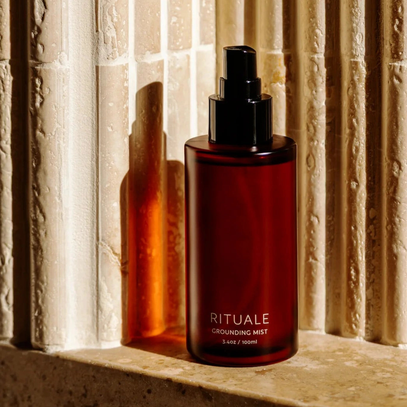 Rituale Grounding Mist amber bottle on stone ledge with warm light and fluted travertine wall background