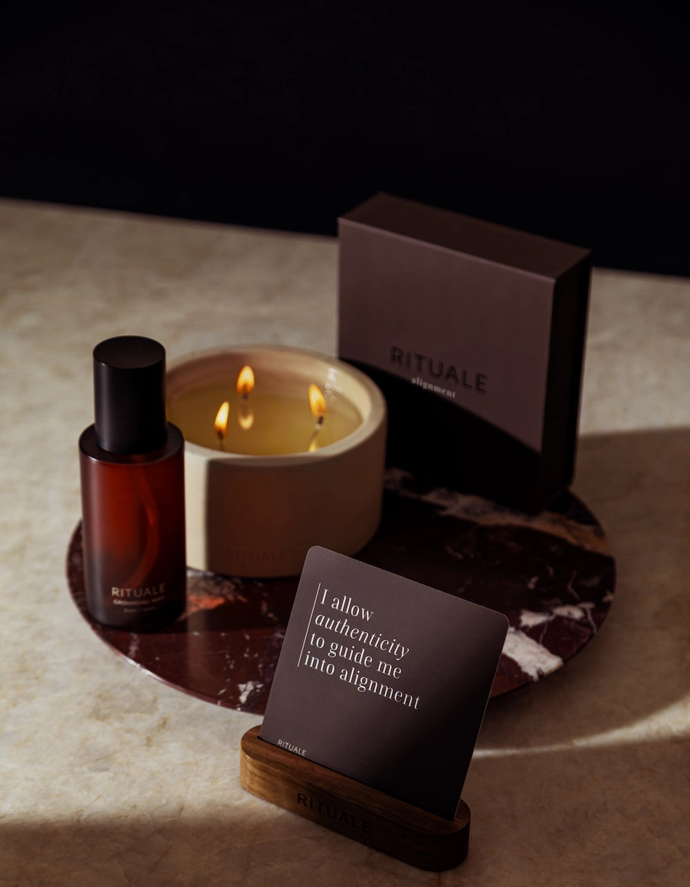 RITUALE grounding mist, concrete candle, and alignment deck affirmation card arranged on a deep red marble tray against a dark background, styled on a cream stone surface with dramatic shadow lighting.